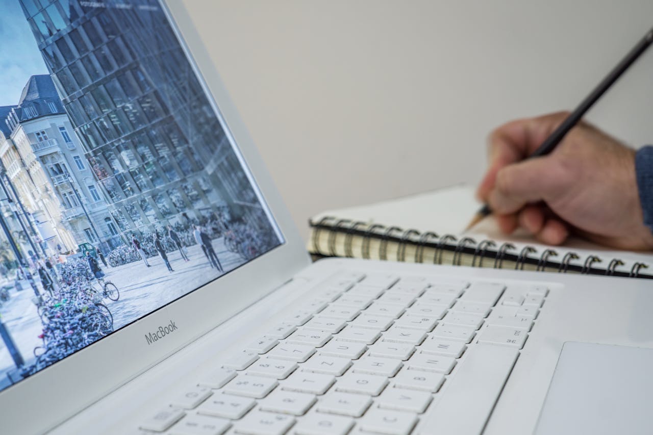 A person taking notes in a notebook beside an open laptop displaying an urban scene.